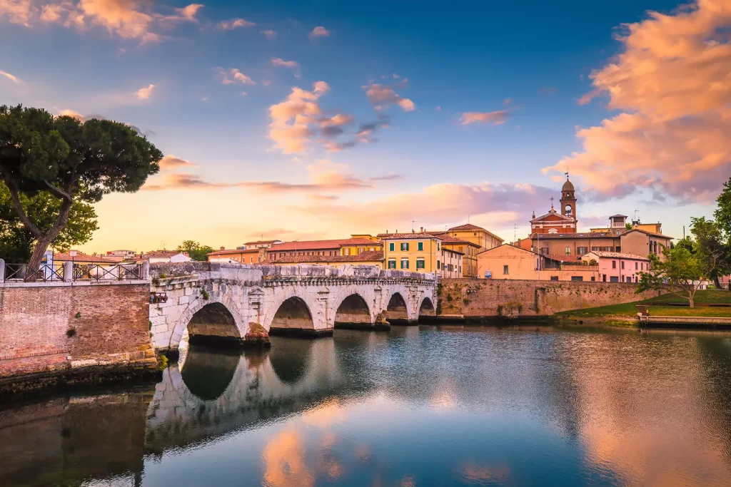 Ponte romano riflesso nell'acqua al tramonto con edifici storici sullo sfondo.