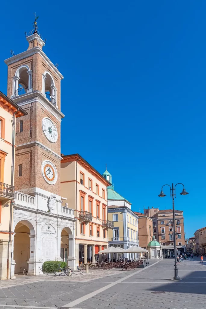 Torre dell'orologio e edifici storici in una piazza soleggiata.