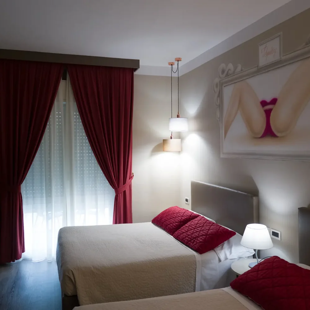 Camera d'albergo moderna con due letti singoli, tende rosse e un grande quadro sopra la testata.
