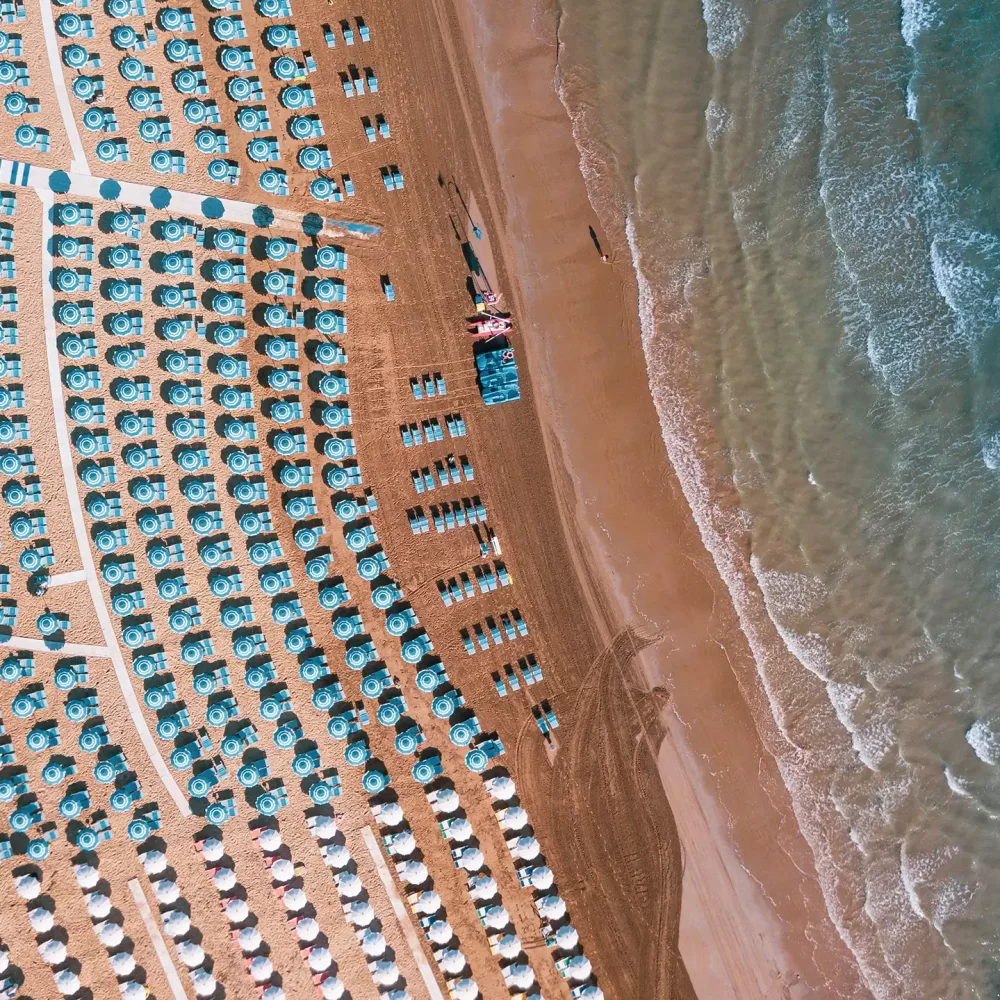 Veduta aerea di una spiaggia affollata con file di ombrelloni e mare ondulato.